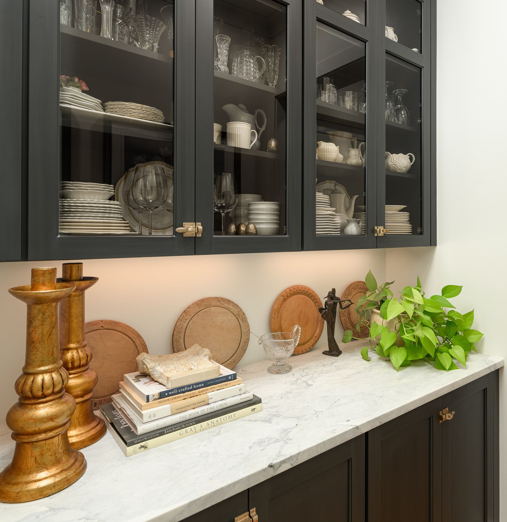 Dark gray kitchen cabinets with glass doors displaying dishware, countertop with décor, and gold candle holders.