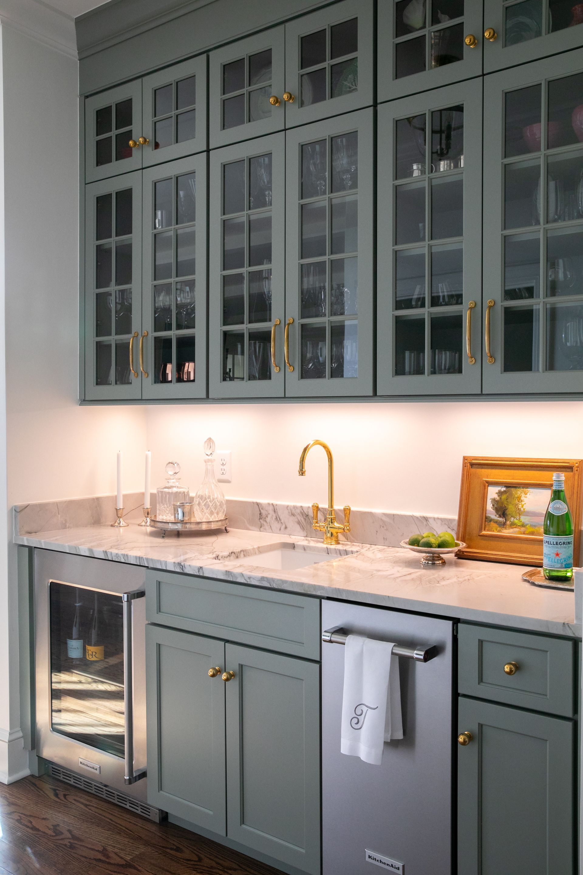 Green built-in bar area with marble countertops, gold hardware, and glass-front cabinets. Includes a sink and mini fridge.