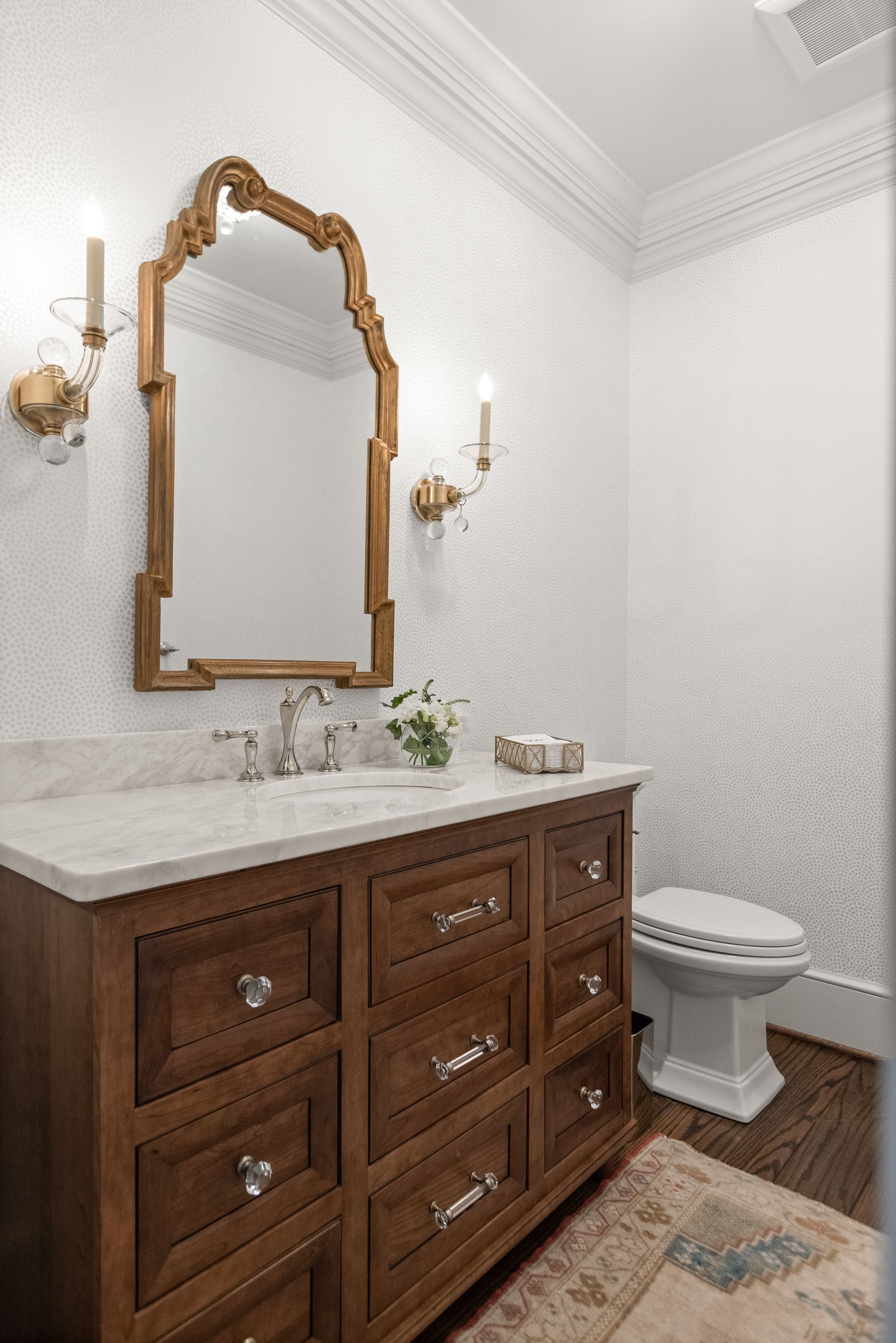 Elegant bathroom with a wooden vanity, ornate mirror, sconces, and patterned wallpaper.