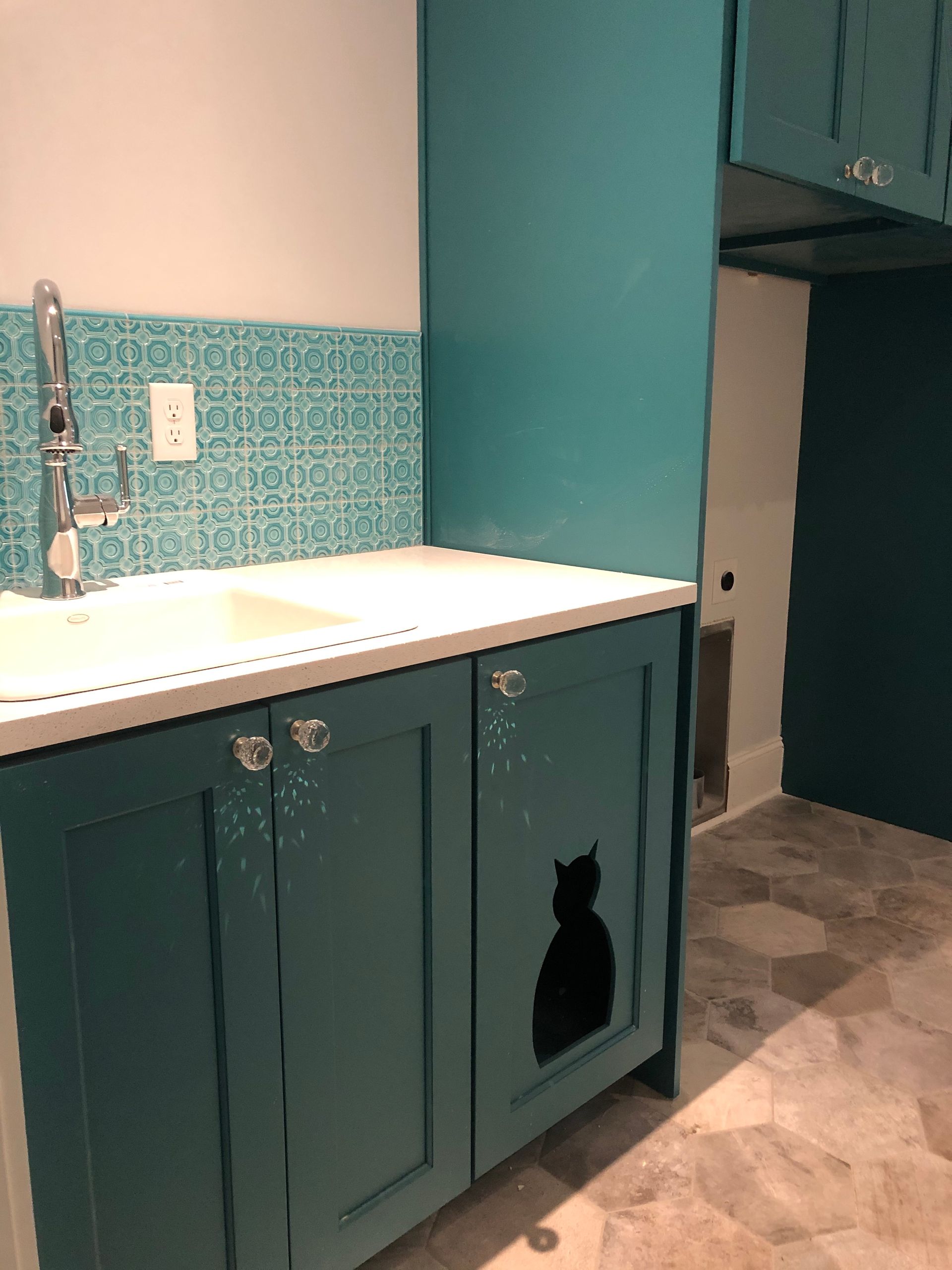 Teal cabinets with cat silhouette, sink with teal backsplash, and doorway to laundry room.