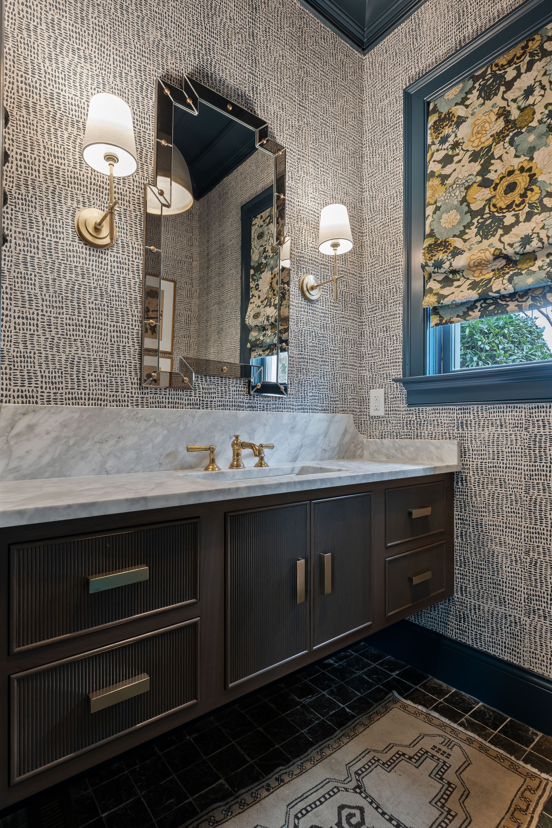 Powder room with patterned wallpaper, dark wood vanity, gold fixtures, and floral window shade.