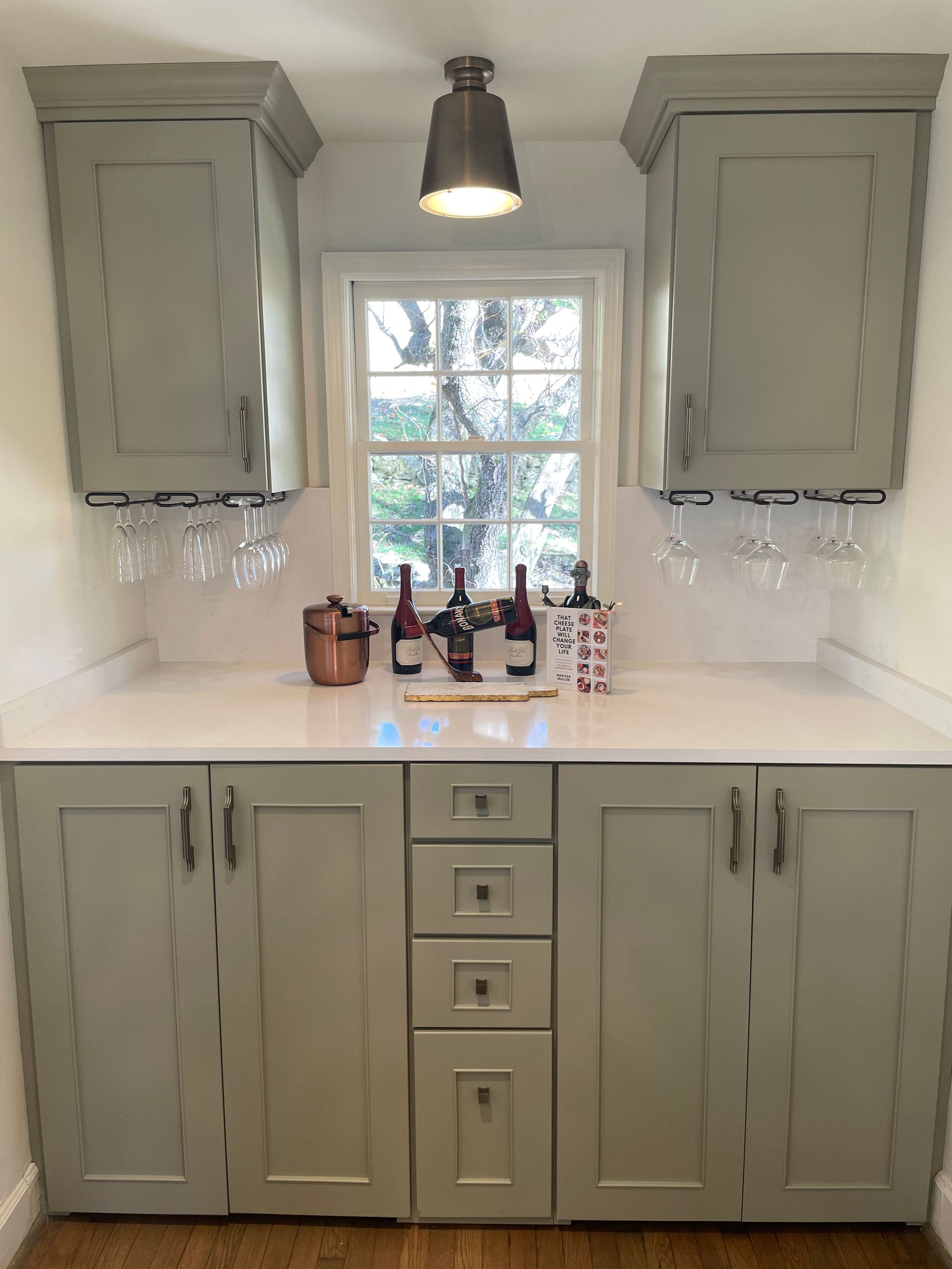 A built-in bar with gray cabinets, a white countertop, a small window, and wine bottles.