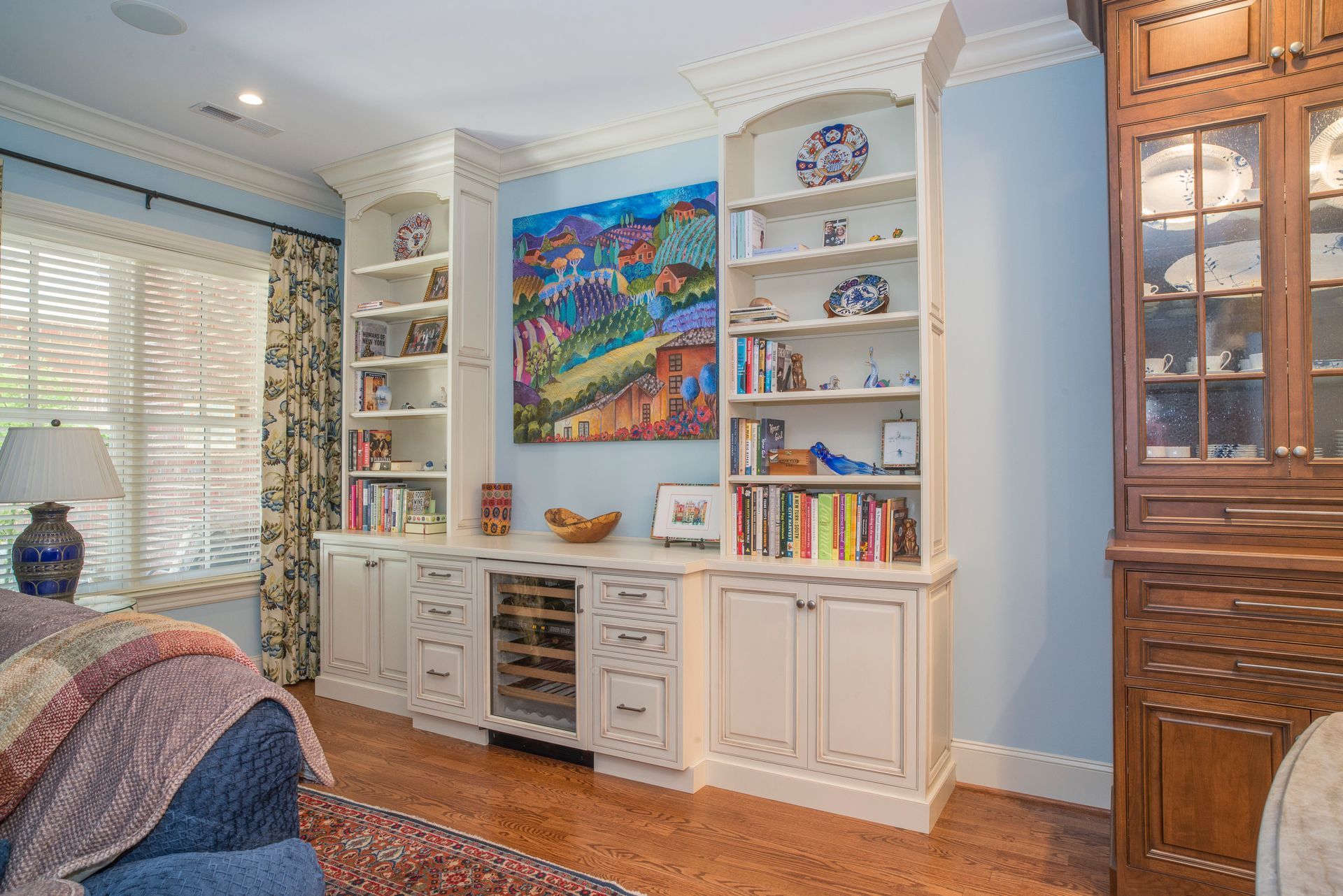 Living room with built-in white shelves, a wine fridge, and colorful artwork on a blue wall, with hardwood floors.