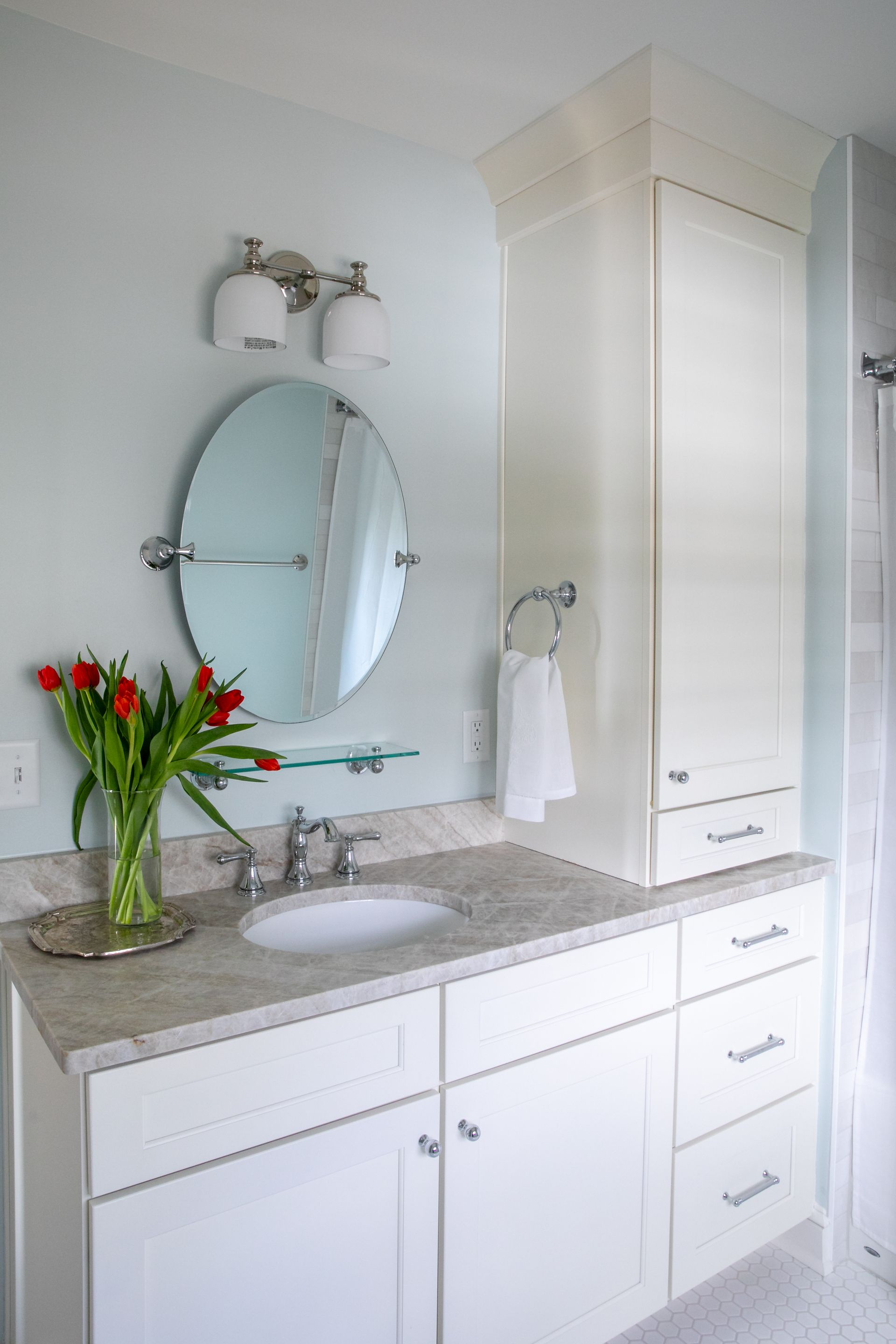 White bathroom with a vanity, oval mirror, and red tulips in a vase.