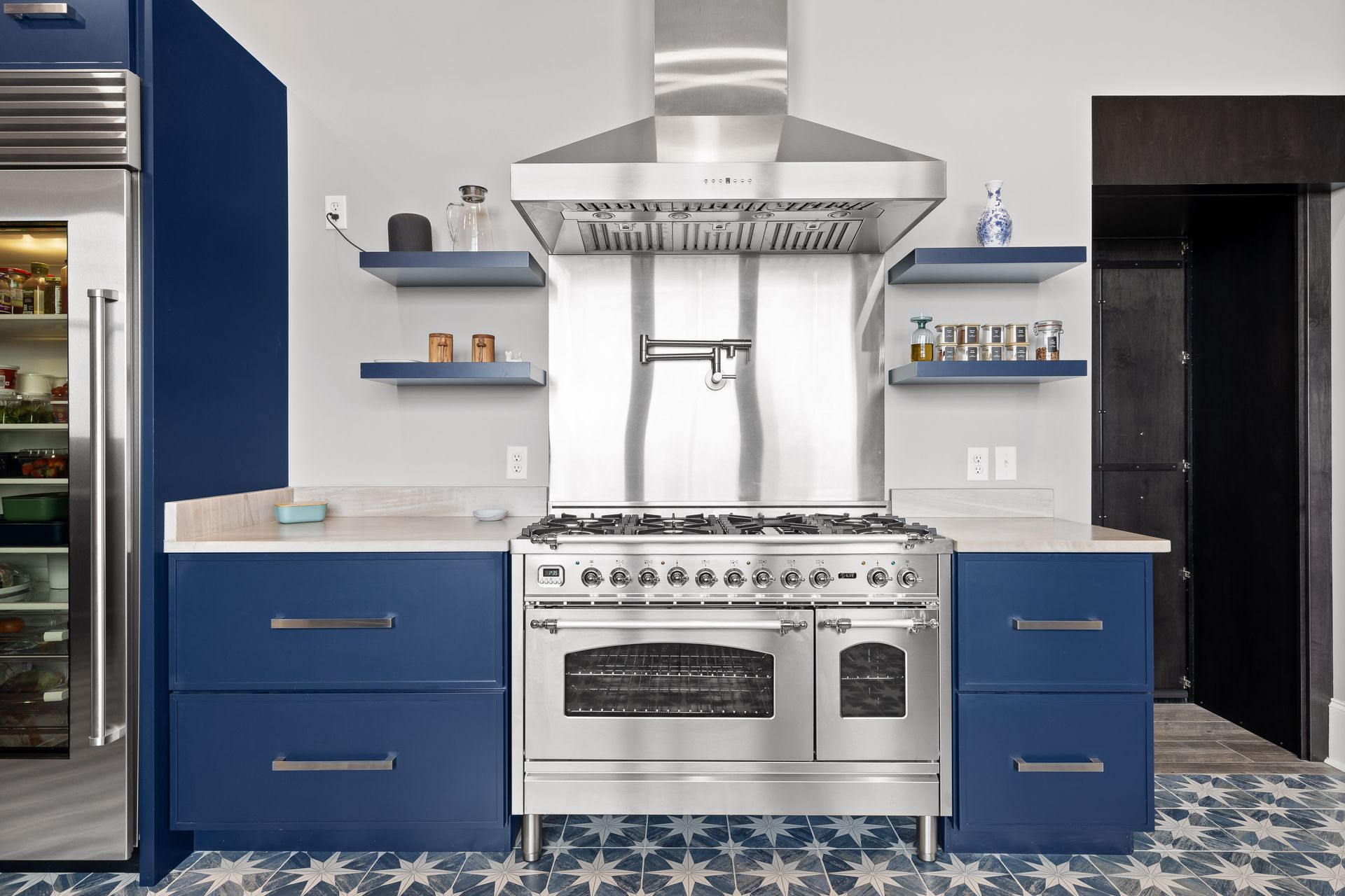 Blue kitchen with stainless steel stove, hood, open shelves, and patterned floor.