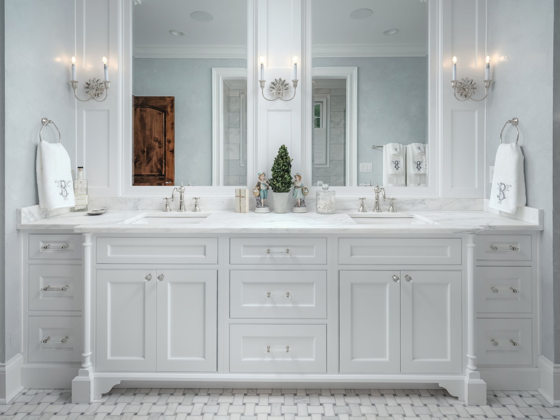 White bathroom vanity with two mirrors, sconces, and a small plant on a white countertop.