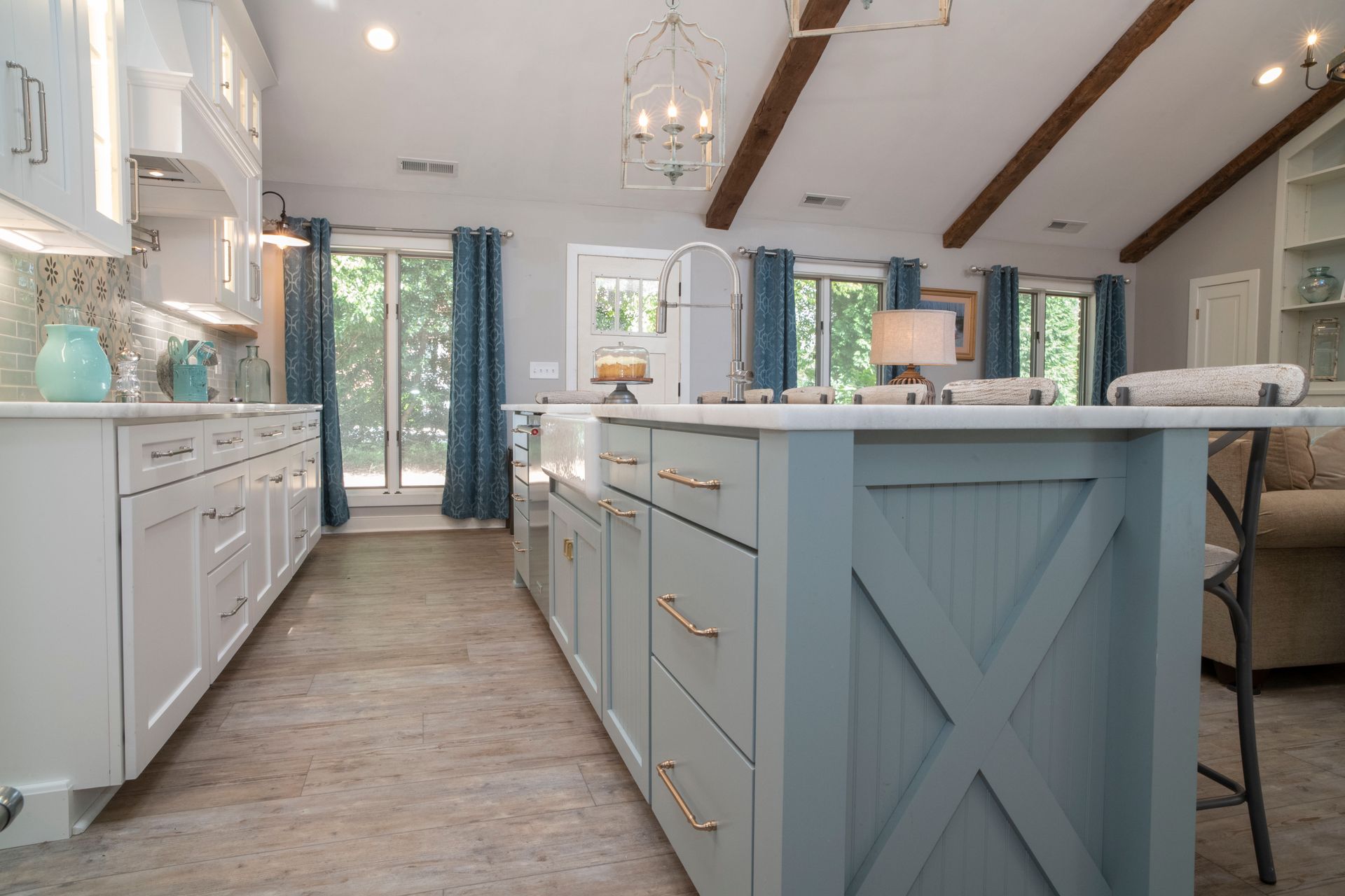 A light-blue farmhouse kitchen with a large island and white cabinets, blue curtains, and a beige floor.
