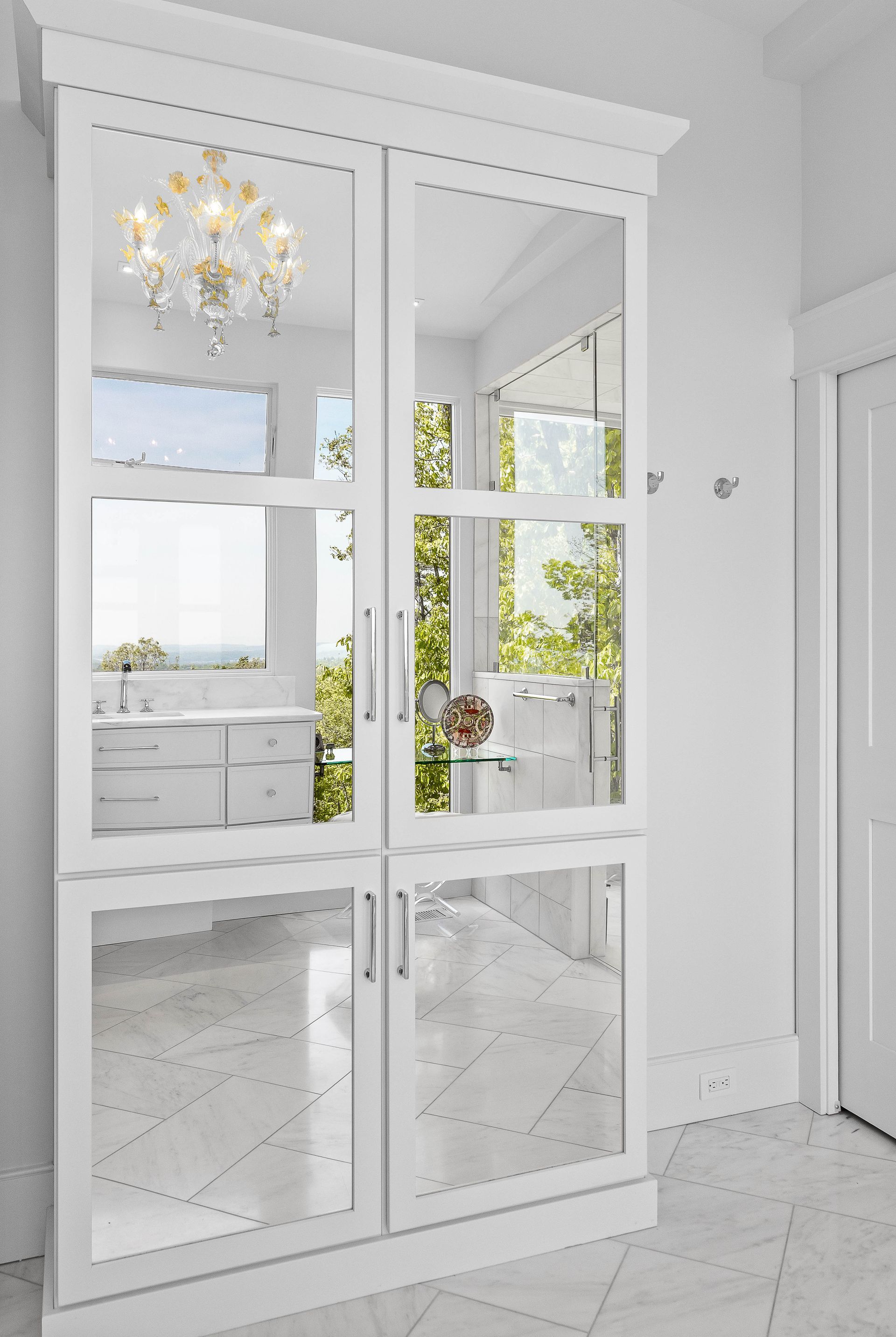 White mirrored cabinet in a hallway, reflecting a bright room with a chandelier and trees.