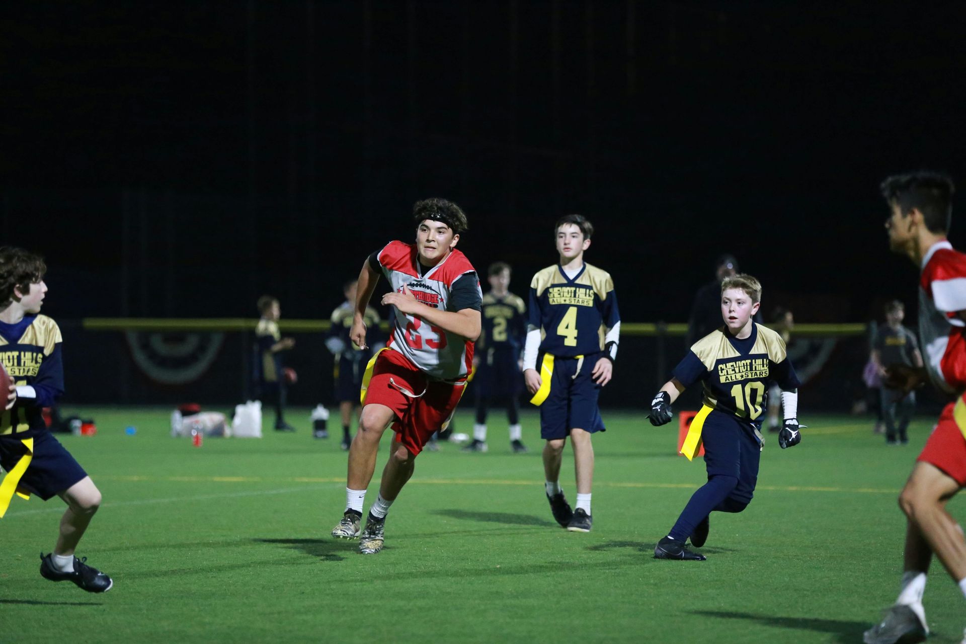 Flag football game at night, a player in red jersey runs with the ball as others pursue on a green field.