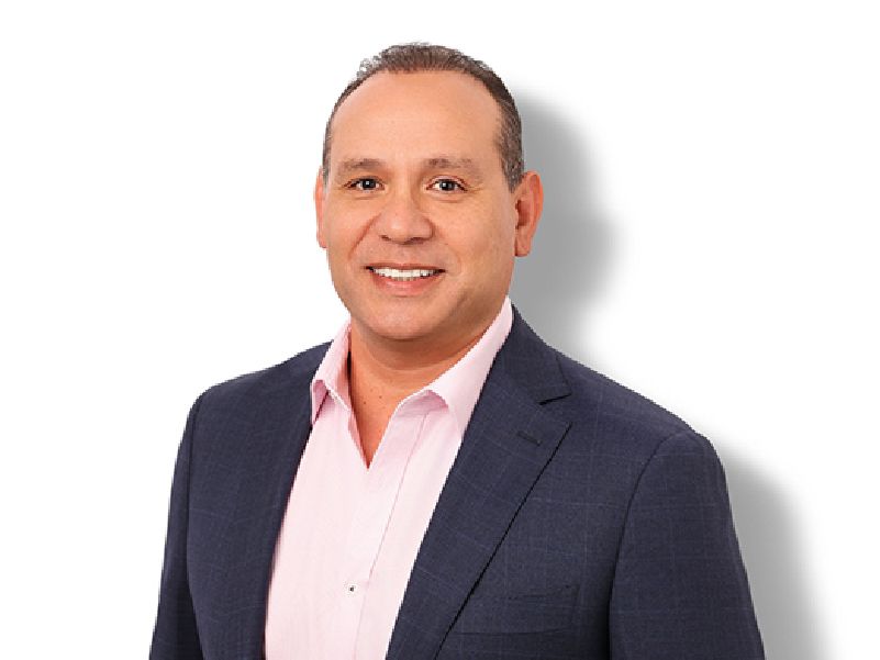 A smiling man with short brown hair wearing a pink shirt and navy blazer.