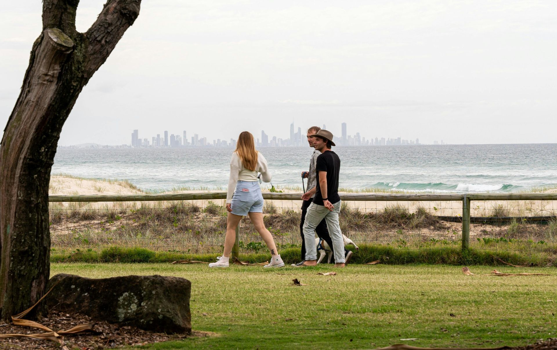 Two people walking on grass near a beach with ocean waves and a distant city skyline