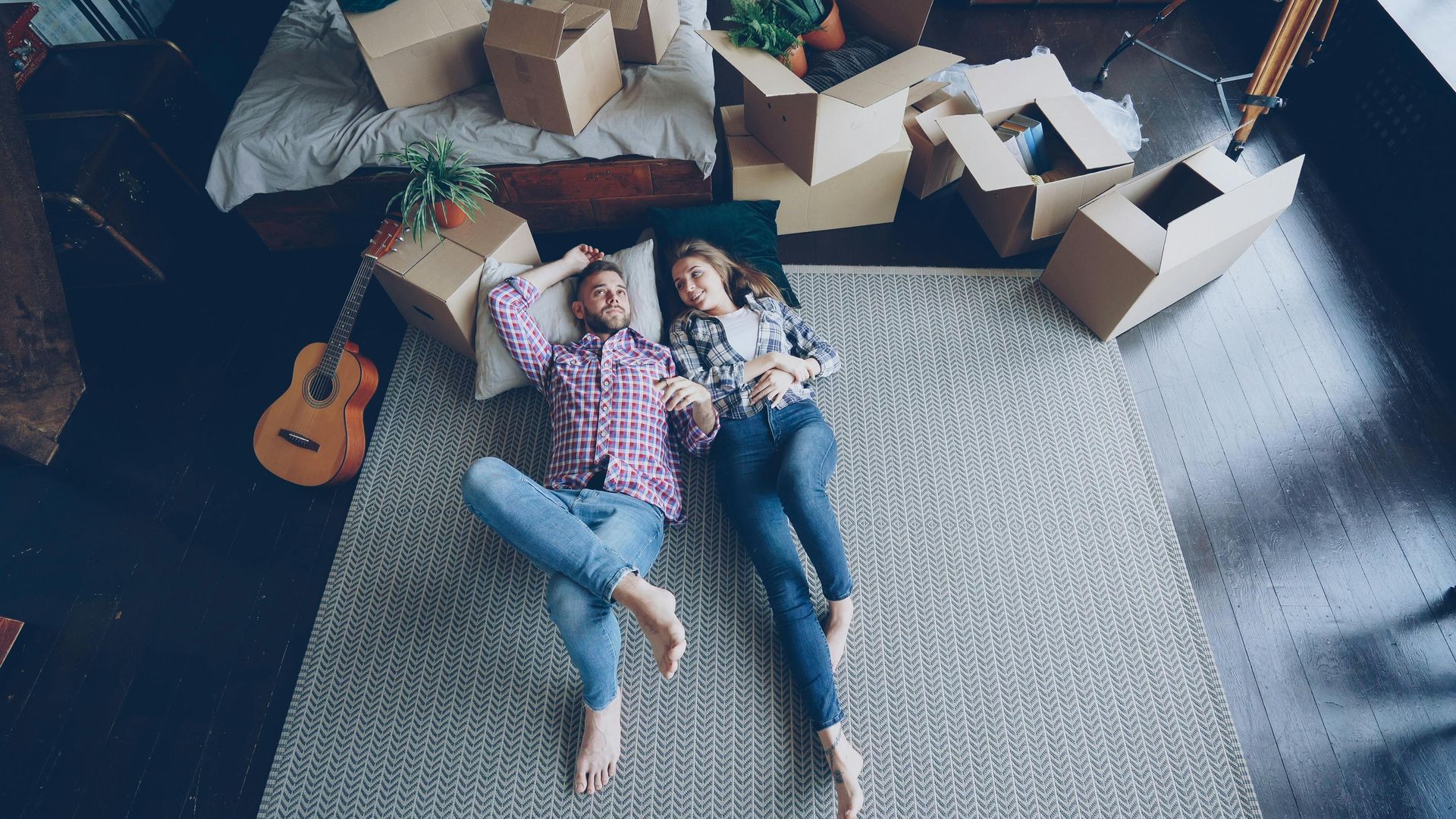 Two people lying on a rug in a room with cardboard boxes, one holding a guitar nearby