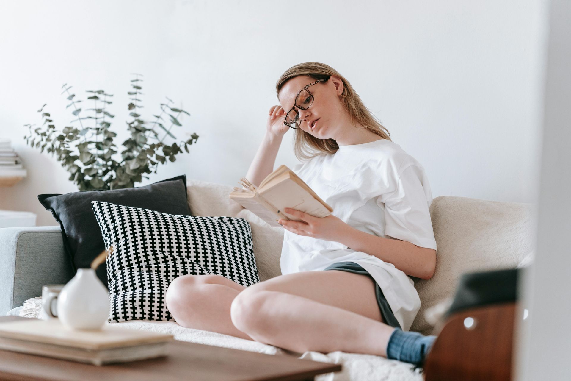 Woman reading a book on a couch in a bright living room with black-and-white pillows.