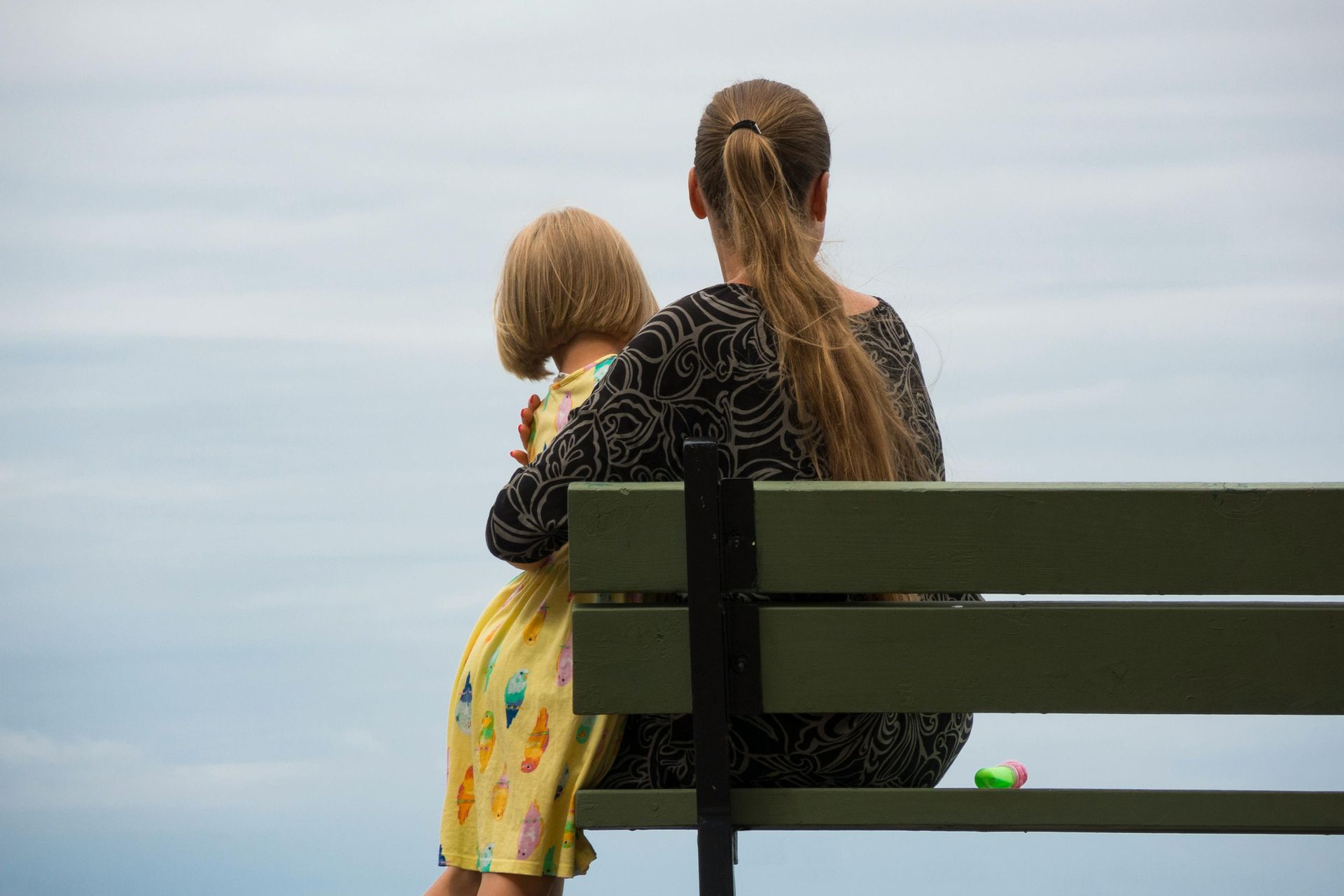 Two children sit on a bench facing a calm sea under a cloudy sky.