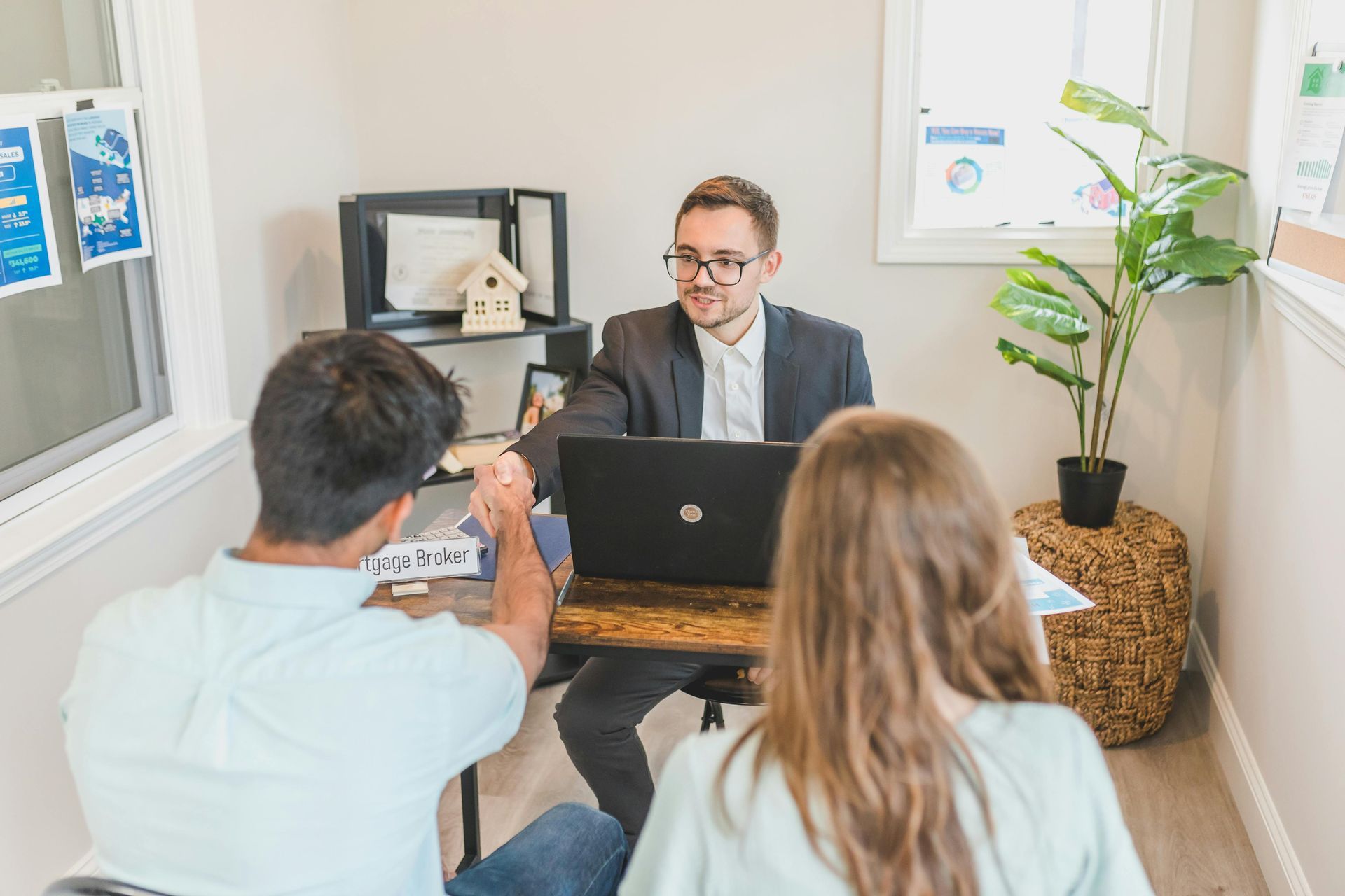 Two people meet with a suited advisor at a desk in a bright office with a laptop and plant.