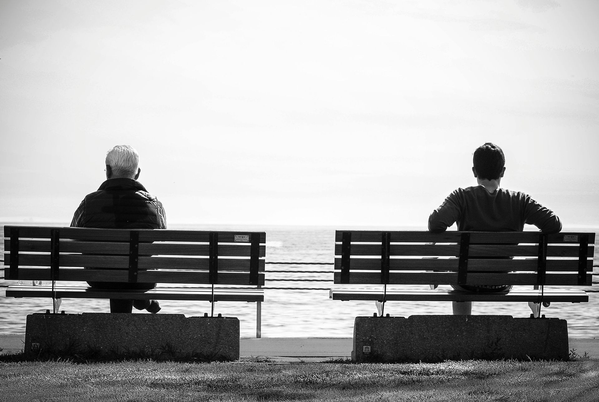 Two people sit on benches facing the ocean, separated by a gap, in a black-and-white scene.