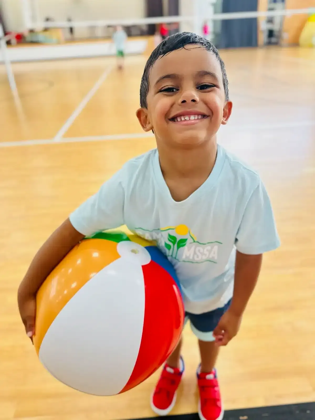 Boy smiling, holding a colorful beach ball in a gymnasium.