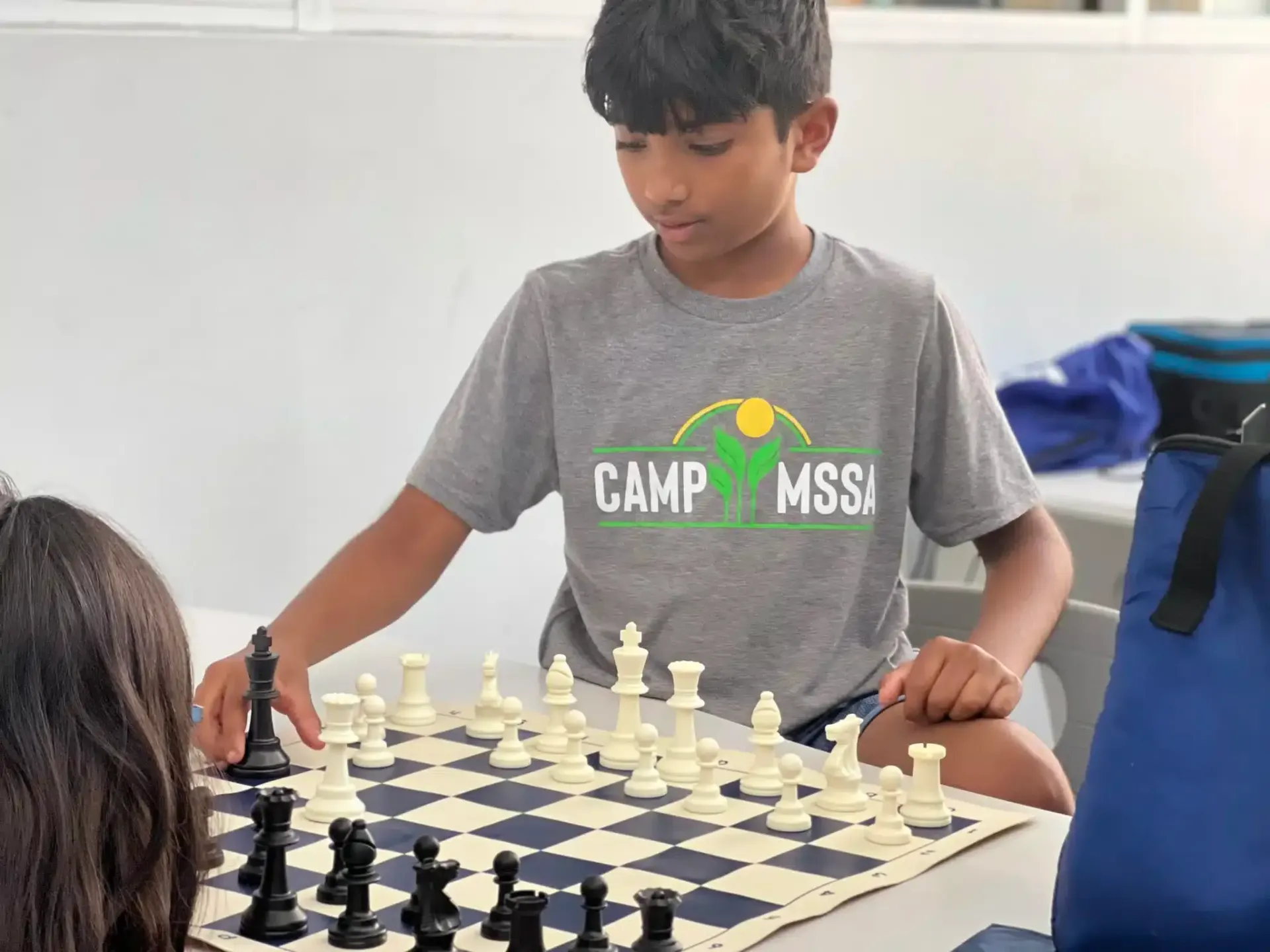 Boy playing chess at a table, looking focused. The board has pieces set up; he's wearing a gray shirt that says Camp MSSA.