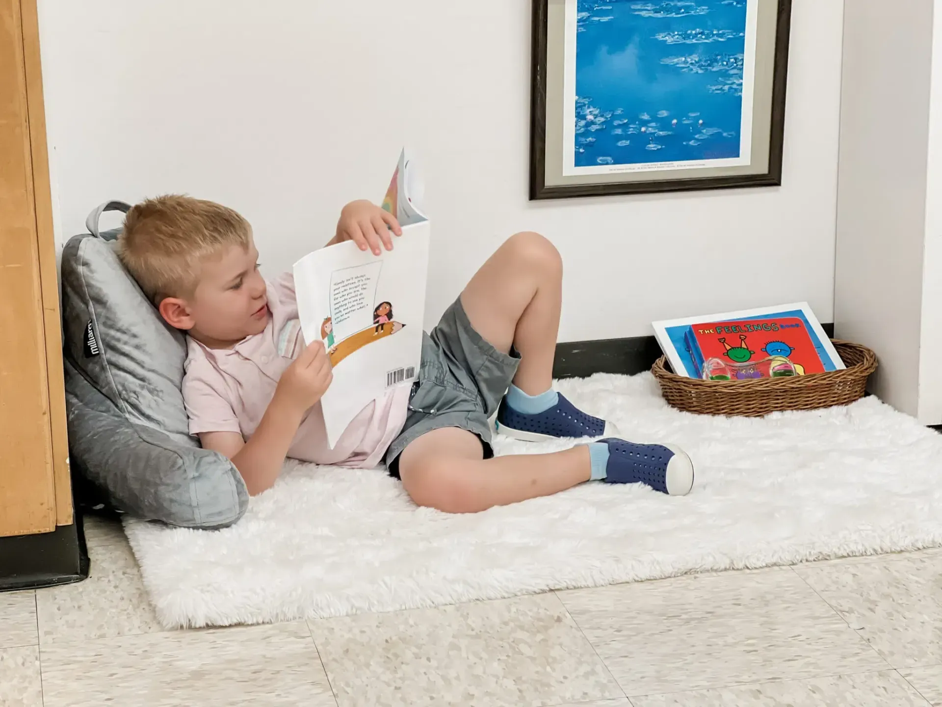 Young child reading a book while lying on a fluffy white rug, near a pillow, wall art, and books in a basket.