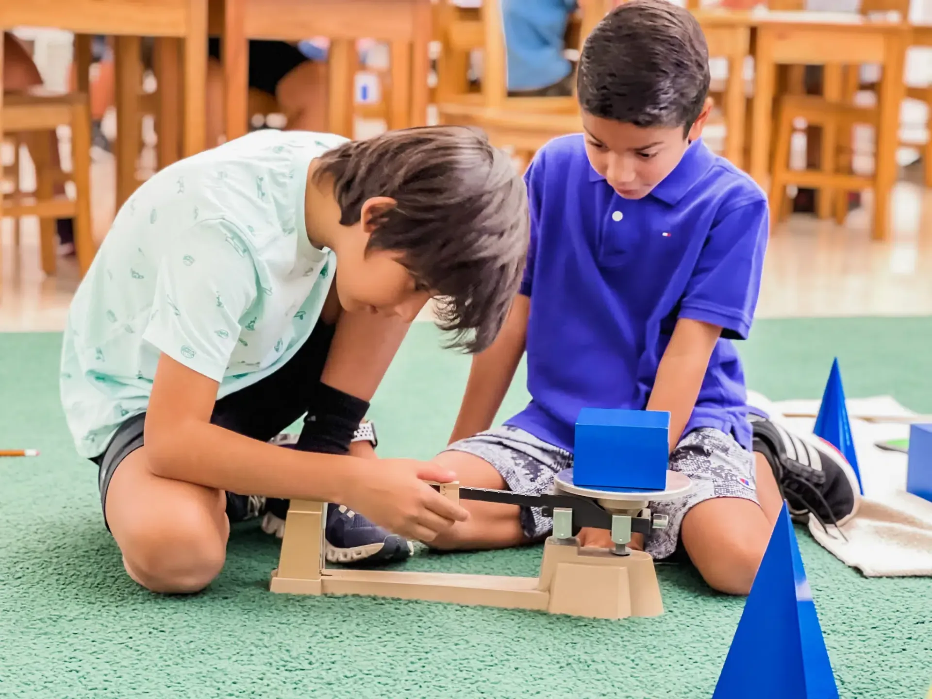 Two boys using a balance scale with a blue cube on a green floor in a classroom.