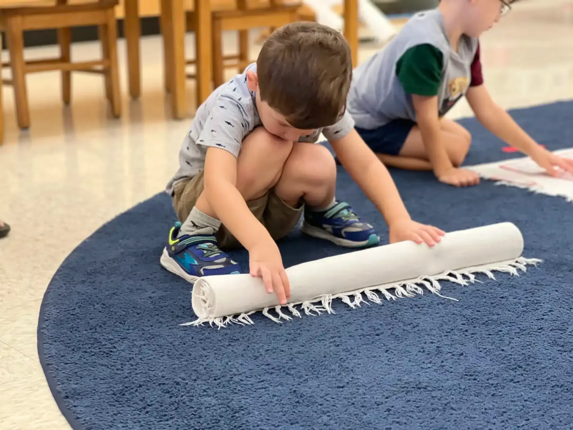 Two children rolling out a white fringed rug on a blue carpet in a room.