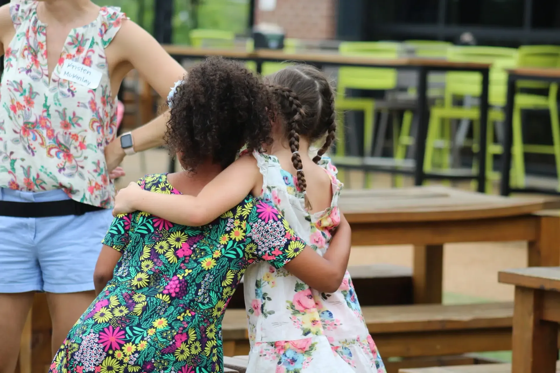 Two girls in floral dresses hugging outside. A woman in floral top and shorts stands near.