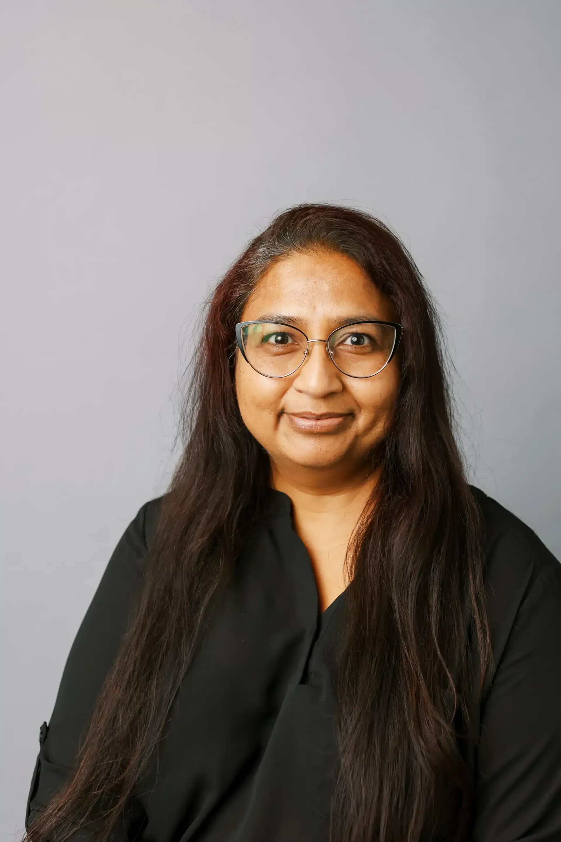 Woman with glasses, long dark hair, and a black shirt smiling, against a gray background.