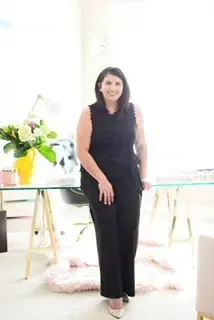 Woman in black sleeveless top and pants smiles, leaning on glass desk in bright office.