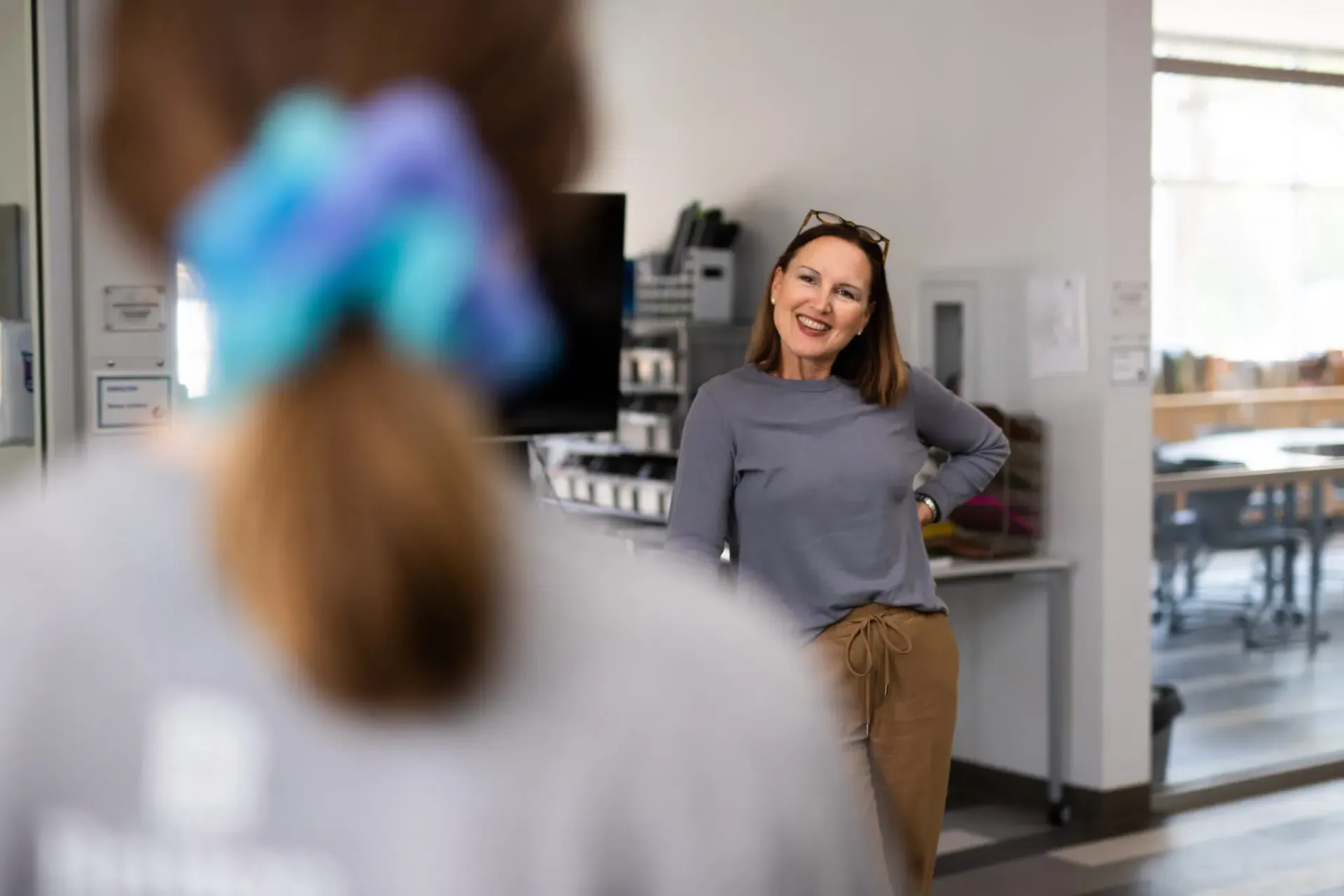 Woman in a grey shirt smiles, looking at someone out of frame; interior shot.