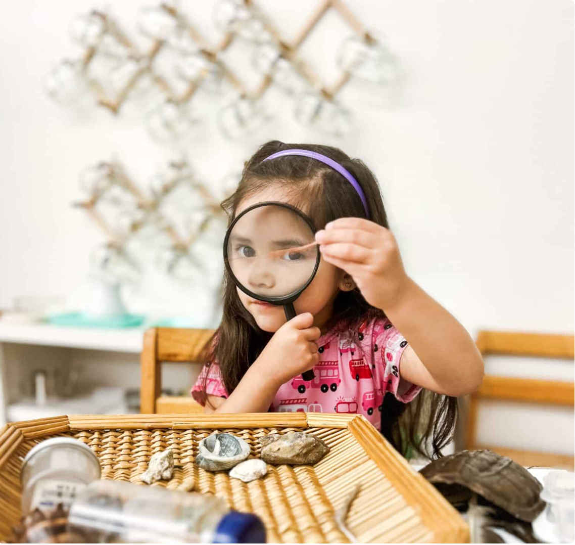 Girl with magnifying glass examining objects on a tray.