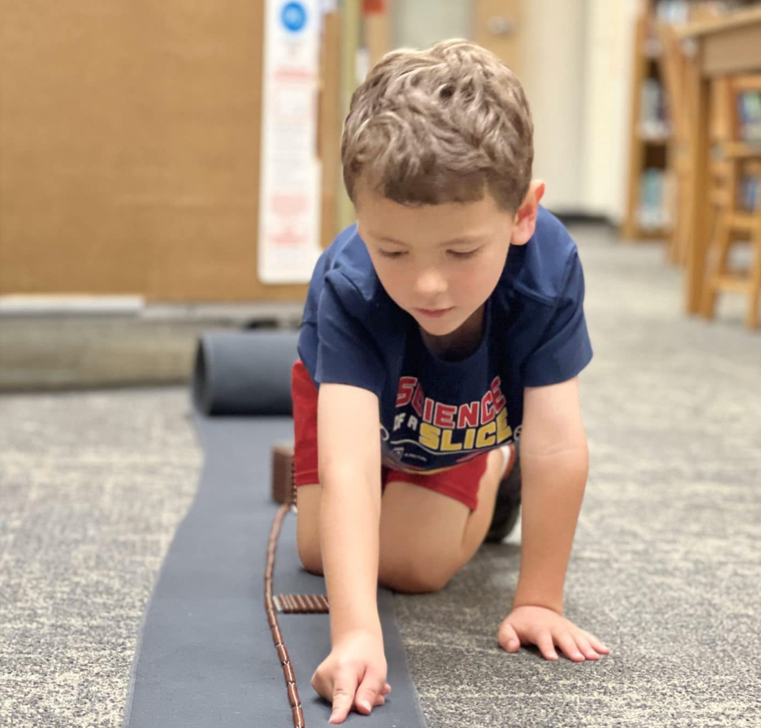 Young child kneeling, arranging dominoes on a gray mat indoors.