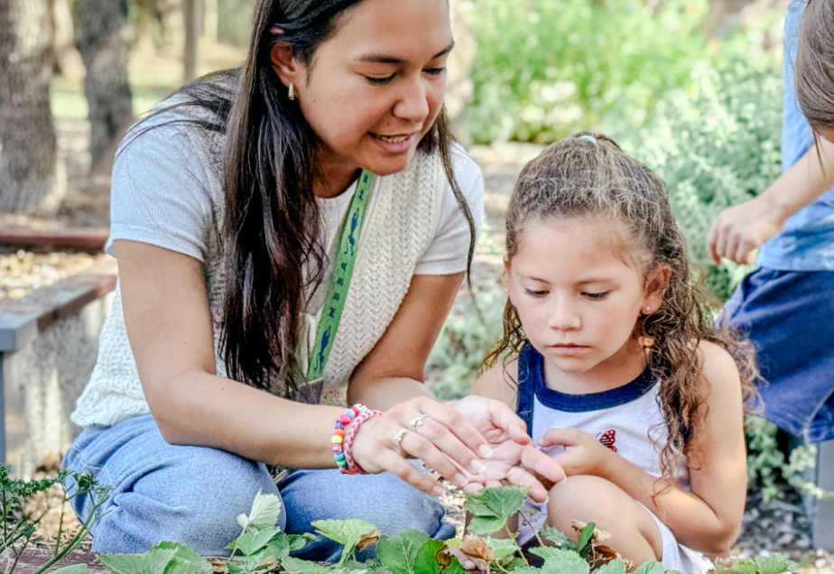 Woman and girl examine leaves outdoors; woman points.