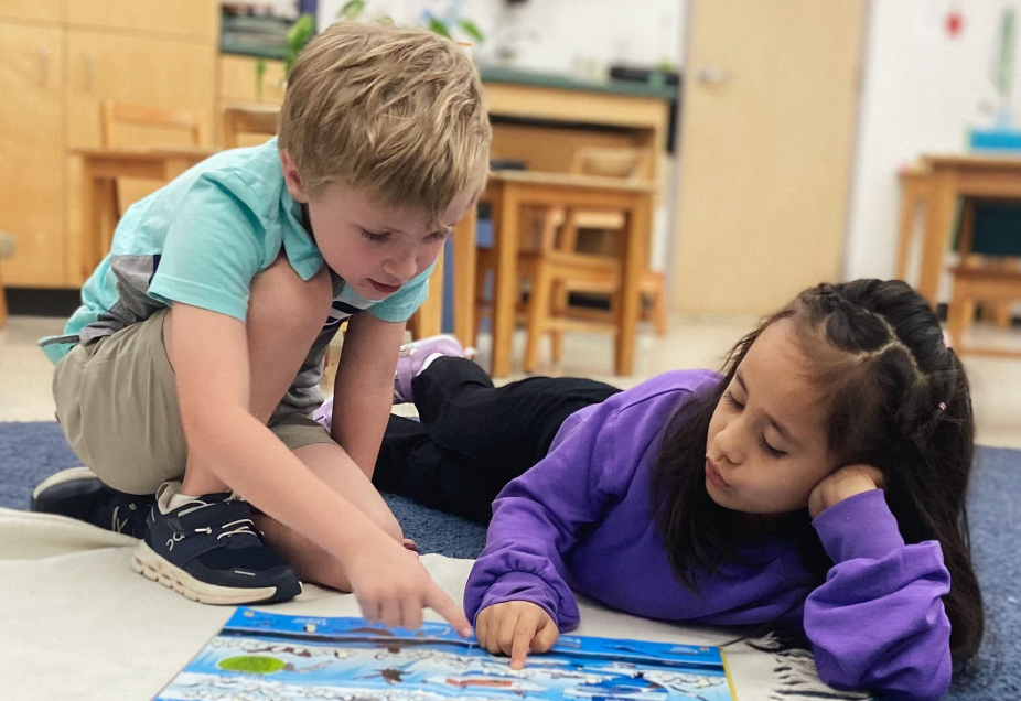 Boy pointing to a book as a girl looks on, both on the floor. Classroom setting.