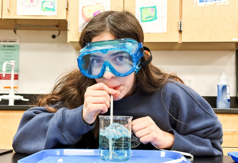 Girl with blue safety goggles drinks from a beaker with a straw in a science lab.