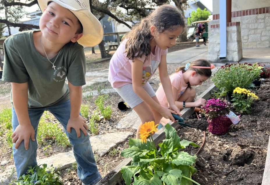 Children planting flowers in a garden bed outdoors; one boy in a cowboy hat smiles.