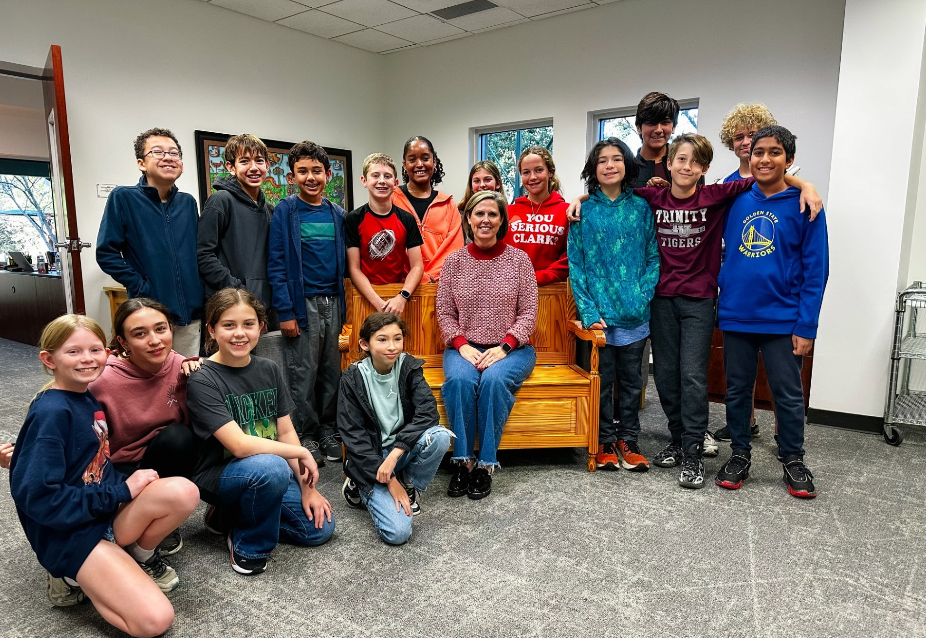 Group of kids and an adult posing together in an office. They smile, near a wooden bench.
