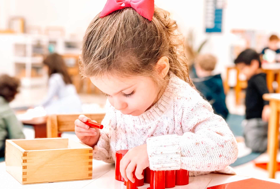 Child with red bow playing with red blocks and wooden box at a table in a classroom.