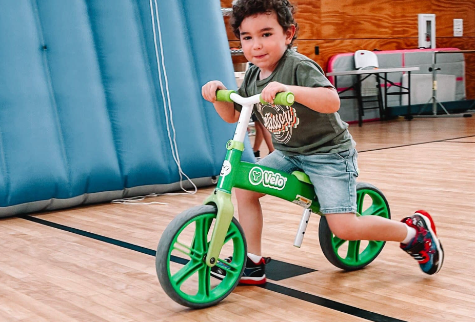 Boy rides a green balance bike inside a gym, smiling at the camera.