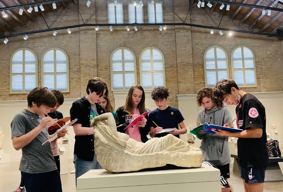 Students writing in notebooks, observing a large sculpture in a museum with arched windows.