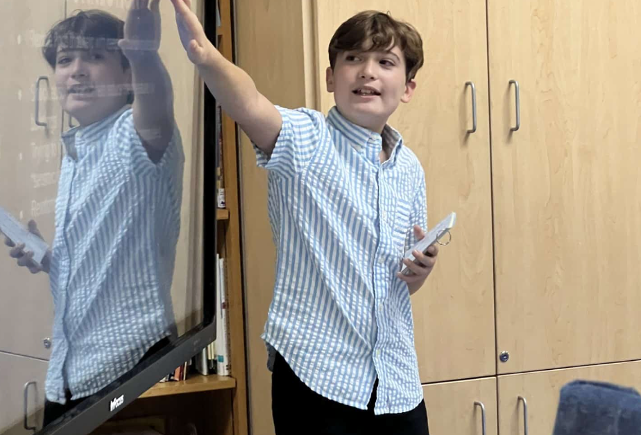 Boy in blue striped shirt gestures by interactive screen, holds paper, standing in front of wooden cabinets.
