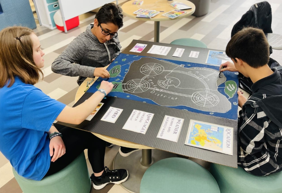 Three students working together on a poster at a table. They are adding finishing touches with markers and are focused on the task.