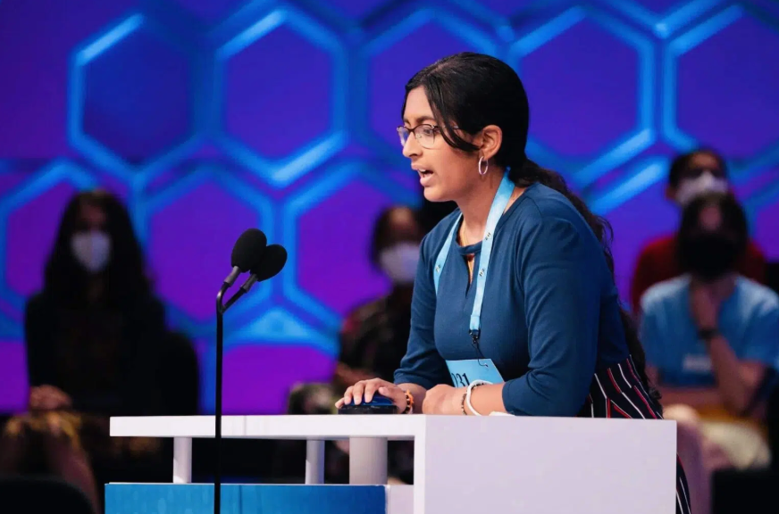A young person spelling at a microphone, wearing glasses and a blue shirt, at a competition with a blue background.