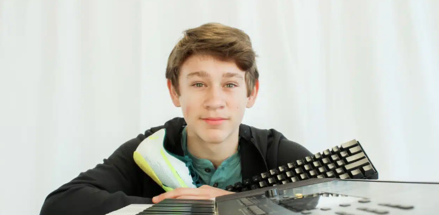 A young person with brown hair smiles, leaning on a keyboard, holding a shoe and keyboard, against a white backdrop.