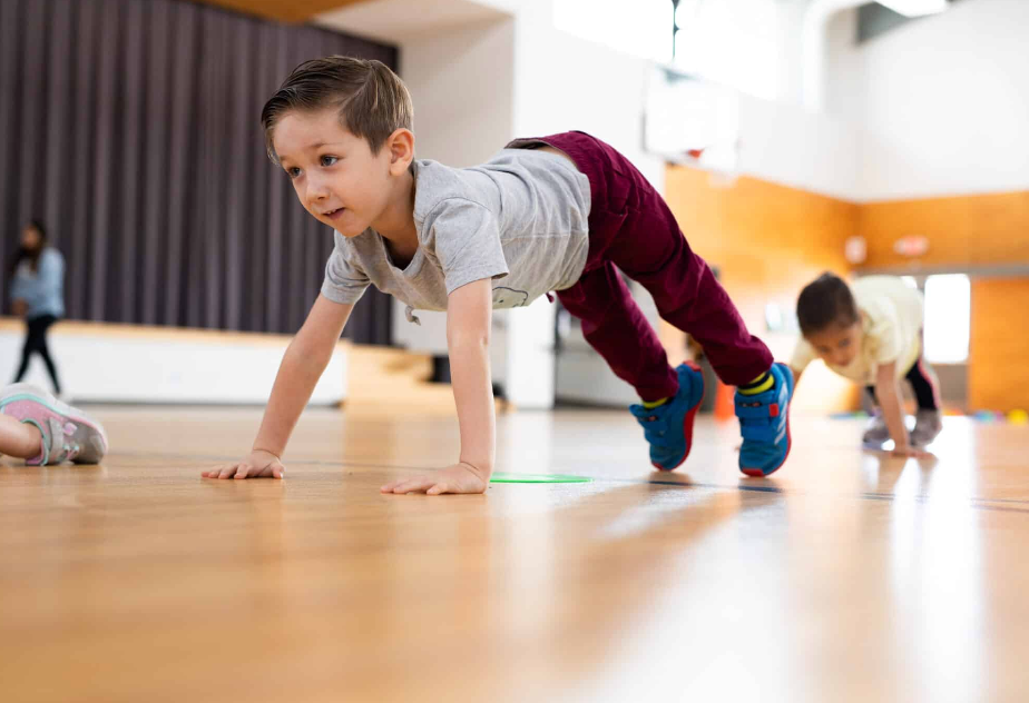 Boy in a gymnasium doing a push-up with another child in the background. Wooden floor, blue shoes, gray shirt.