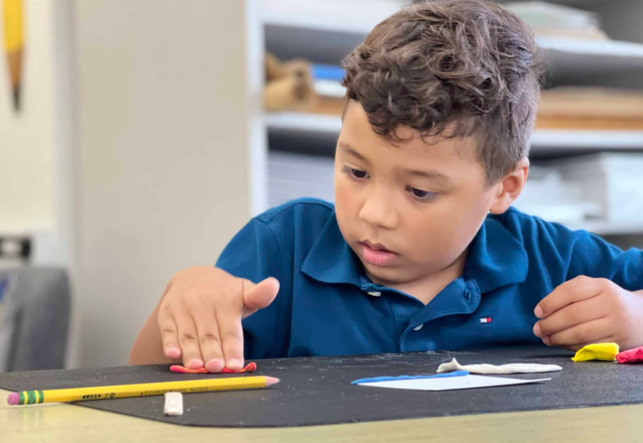 Boy focused on flattening clay at a table, pencil and eraser nearby.