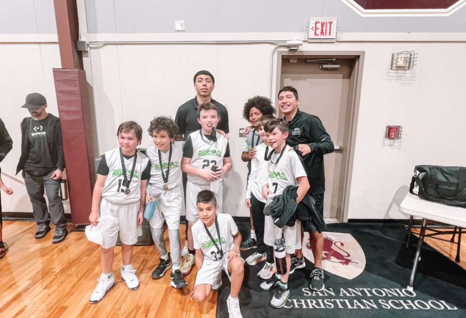 Basketball team poses with coach after game, smiling. Team is wearing matching jerseys with medals. San Antonio Christian School logo visible.
