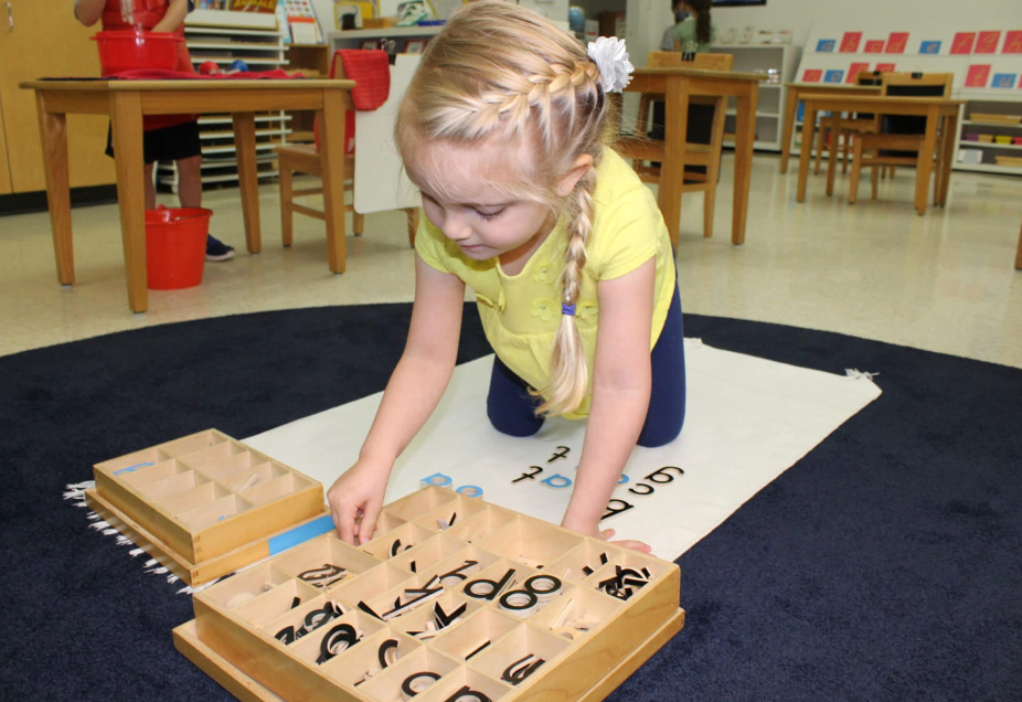 A child on her knees, working with wooden number trays on a mat in a classroom setting.