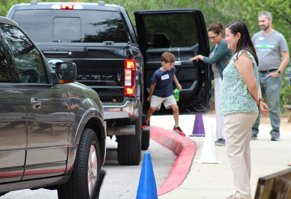 A boy exits a black truck, stepping onto a red curb with help from adults. Blue and white traffic cones are nearby.