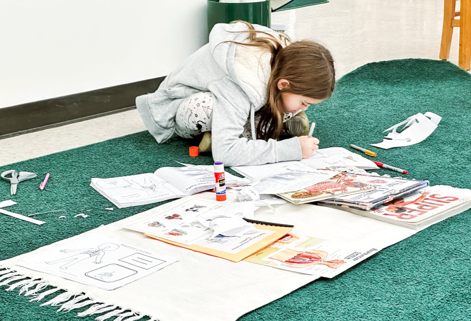 Girl, crouched on green carpet, drawing in coloring books, surrounded by art supplies.