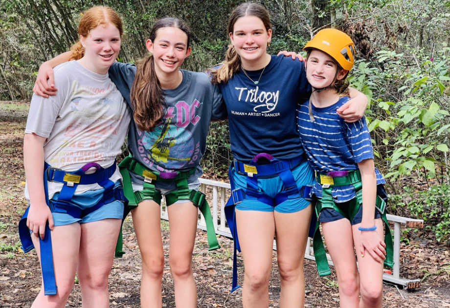 Four people in safety harnesses pose outdoors, smiling. One wears a helmet.